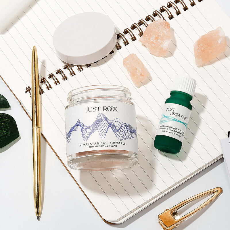 Just Breathe Essential Oils Blend Aromatherapy set from Elan Skincare placed on a notebook , Himalayan salt crystals scattered around the open jar and the green Just Breathe bottle, pen, desk. Diffuser oils scents.